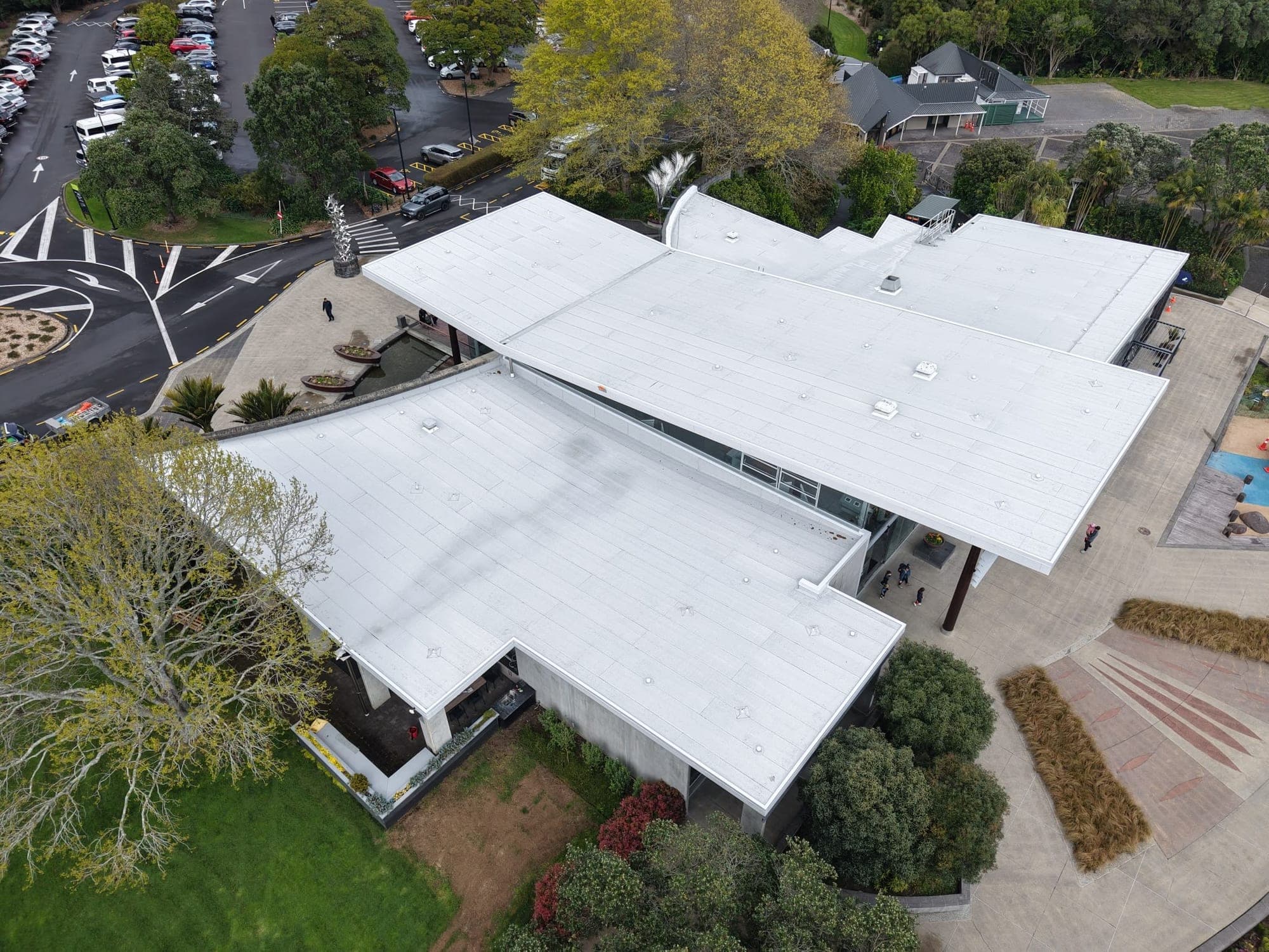 Angled drone view across the finished white membrane roofs of the Visitors Centre, with the forecourt on one side and the carpark beyond